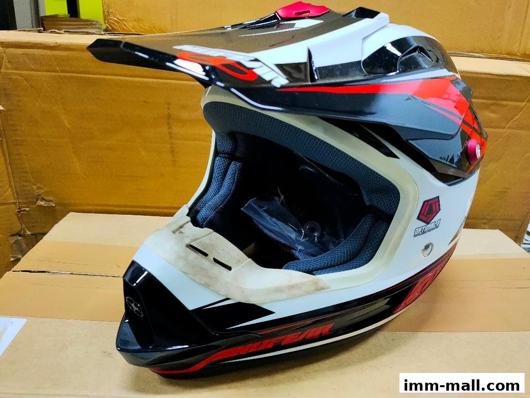 A white, black, and red motocross helmet with a visor is displayed on a cardboard surface.