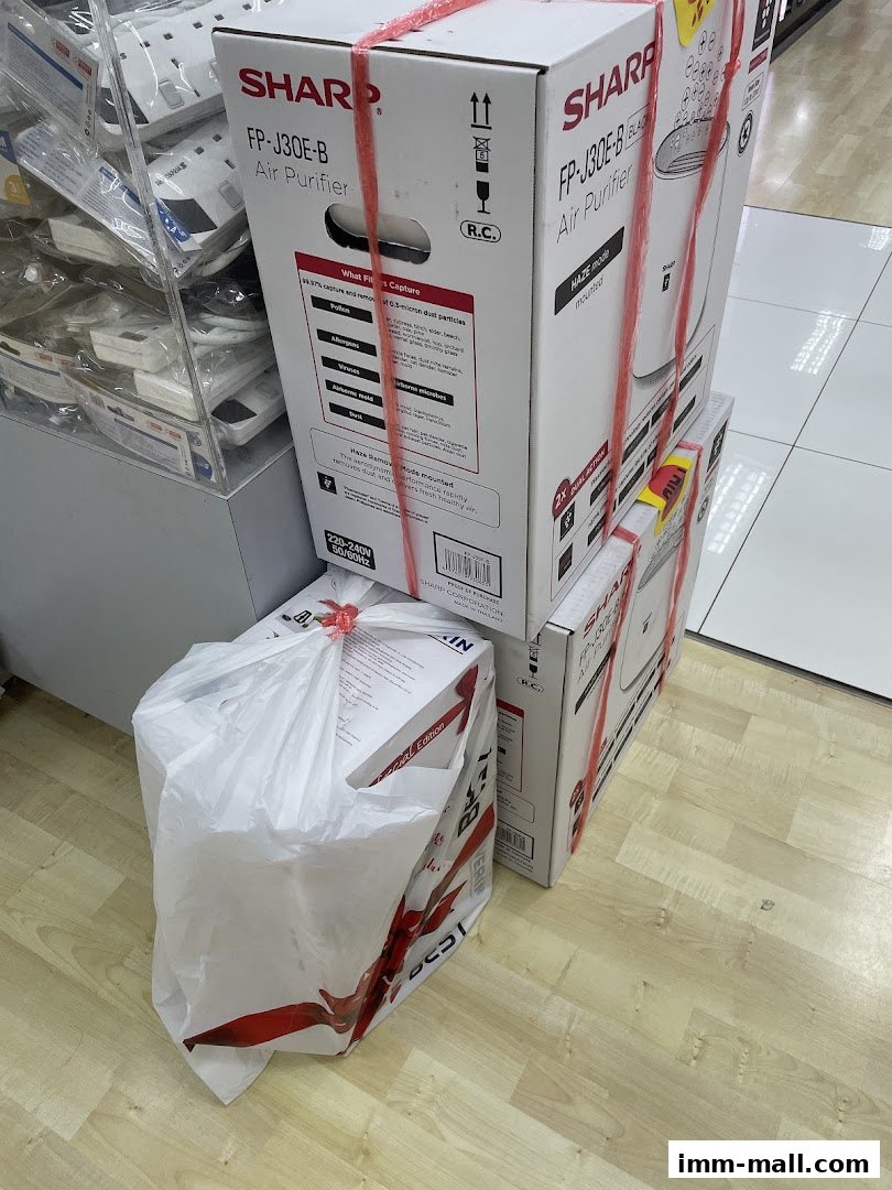Several Sharp air purifiers are stacked and secured with red ties, with one in a Best Denki shopping bag.