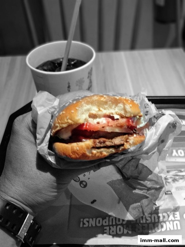 A hand holds a partially eaten Whopper with a drink in the background.