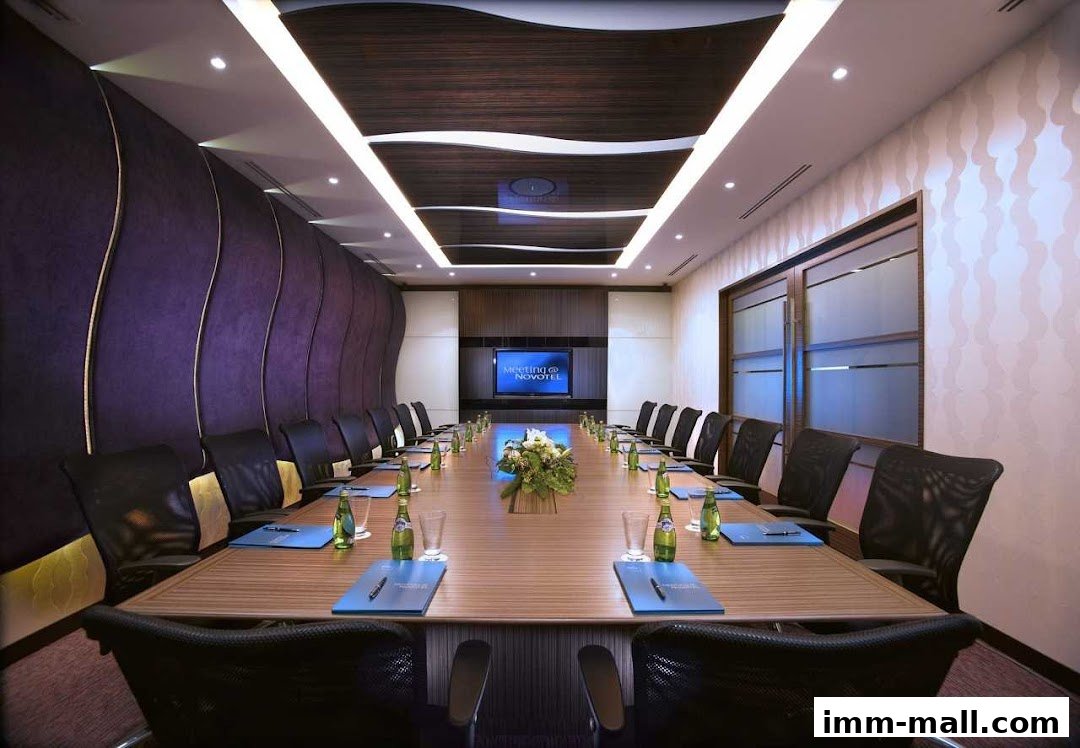 A modern conference room with a long table, chairs, and a screen displaying 'MEETING @ NOVOTEL'.