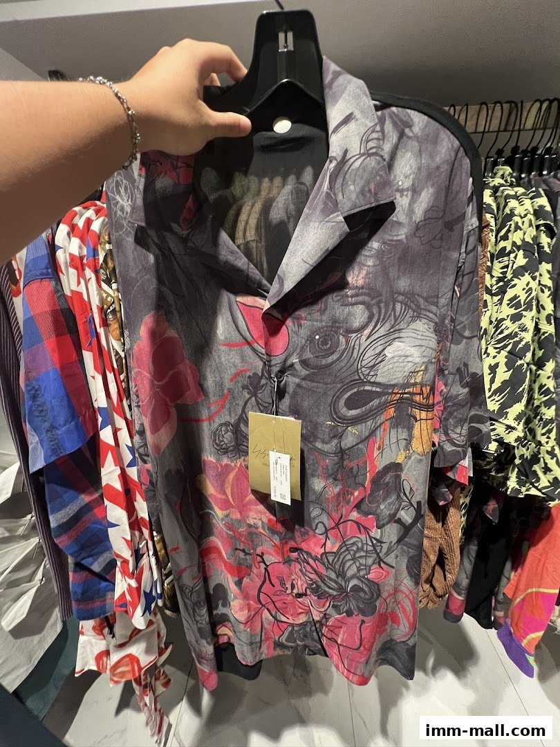 A person holds up a stylish, patterned shirt from Club 21, surrounded by other clothing.