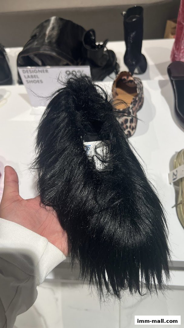 A hand holds a black furry shoe in front of a display of designer shoes.