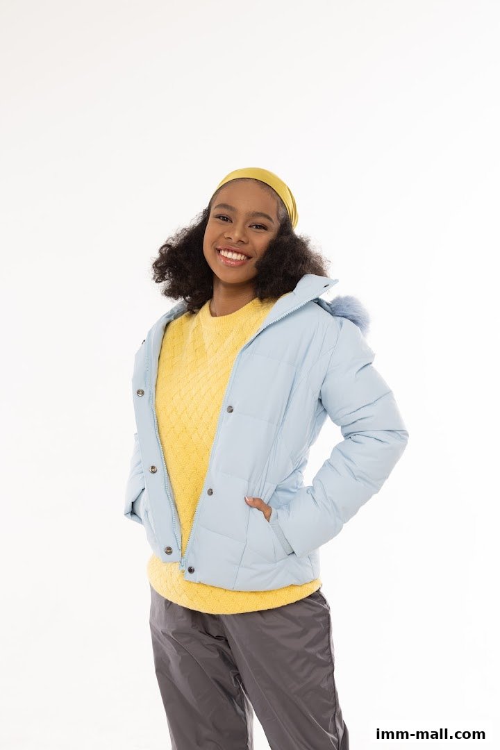 A smiling young woman wearing a light blue puffer jacket, yellow sweater, and grey pants.