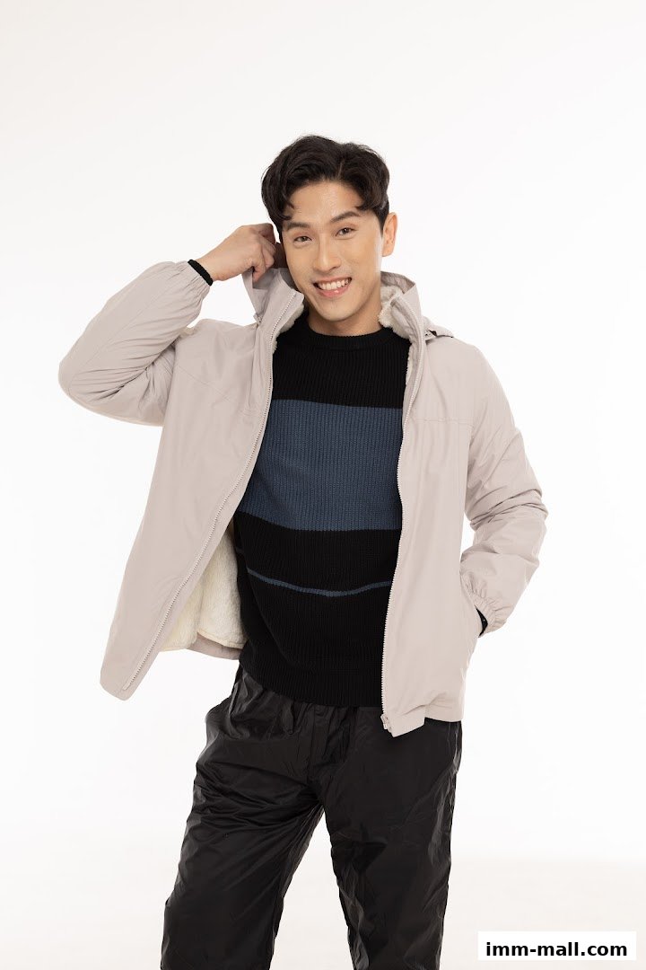A smiling man wearing a light-colored jacket, striped sweater, and black pants.