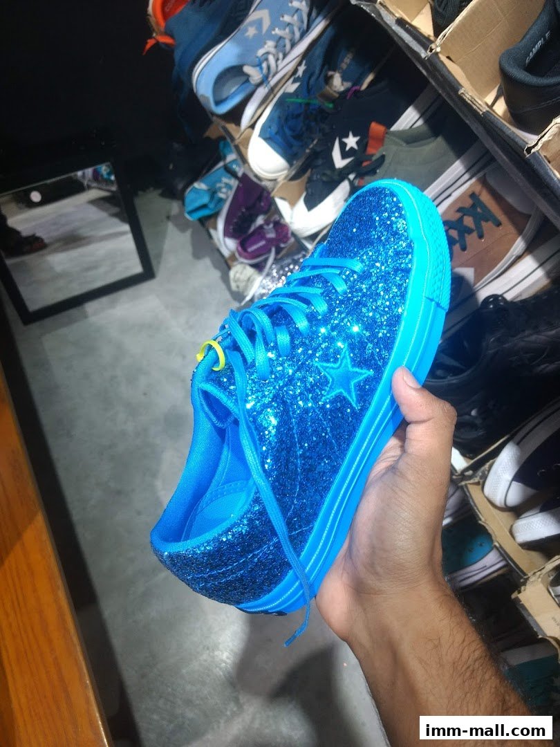 A hand holds a sparkly blue Converse sneaker with other shoes in the background.