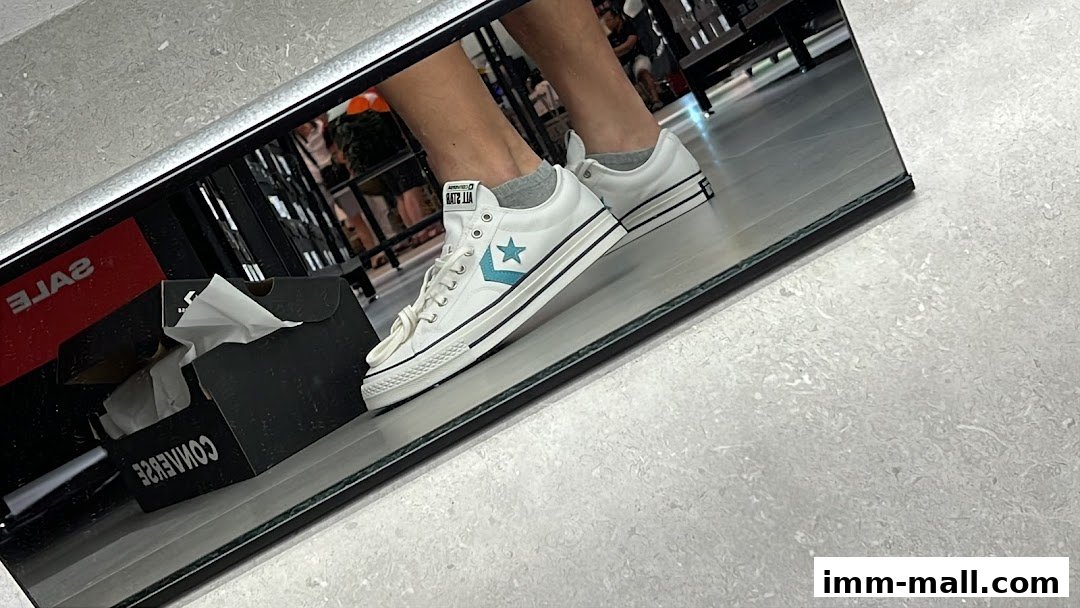 A person tries on white Converse sneakers reflected in a mirror with a sale sign.
