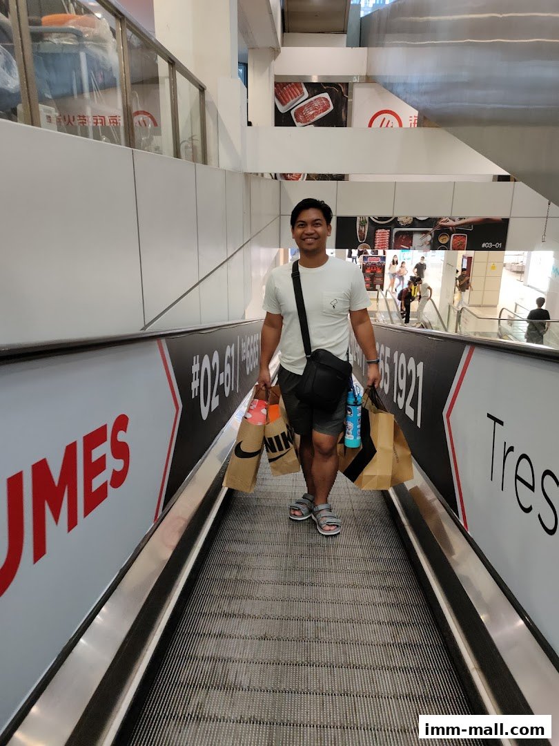 A man smiles while holding shopping bags on an escalator in a mall.