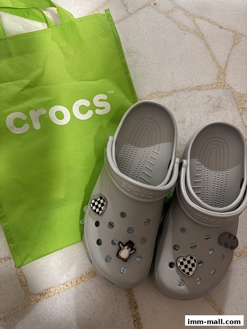 A pair of grey Crocs with Jibbitz charms and a green Crocs shopping bag.