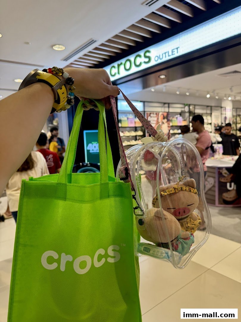 A person holds a green Crocs shopping bag and a clear bag with plush toys in front of a Crocs Outlet store.