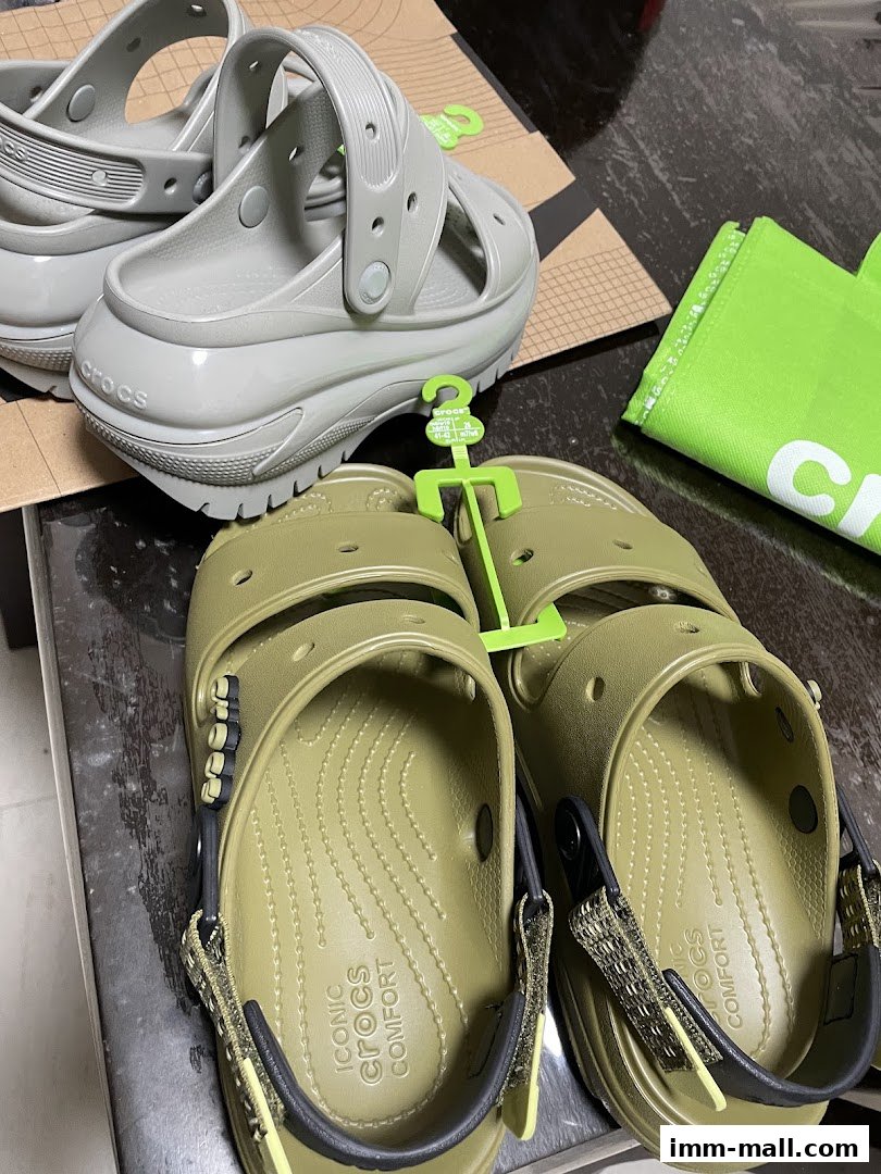 A close-up view of two pairs of Crocs shoes, one grey and one olive green, with a green shopping bag.