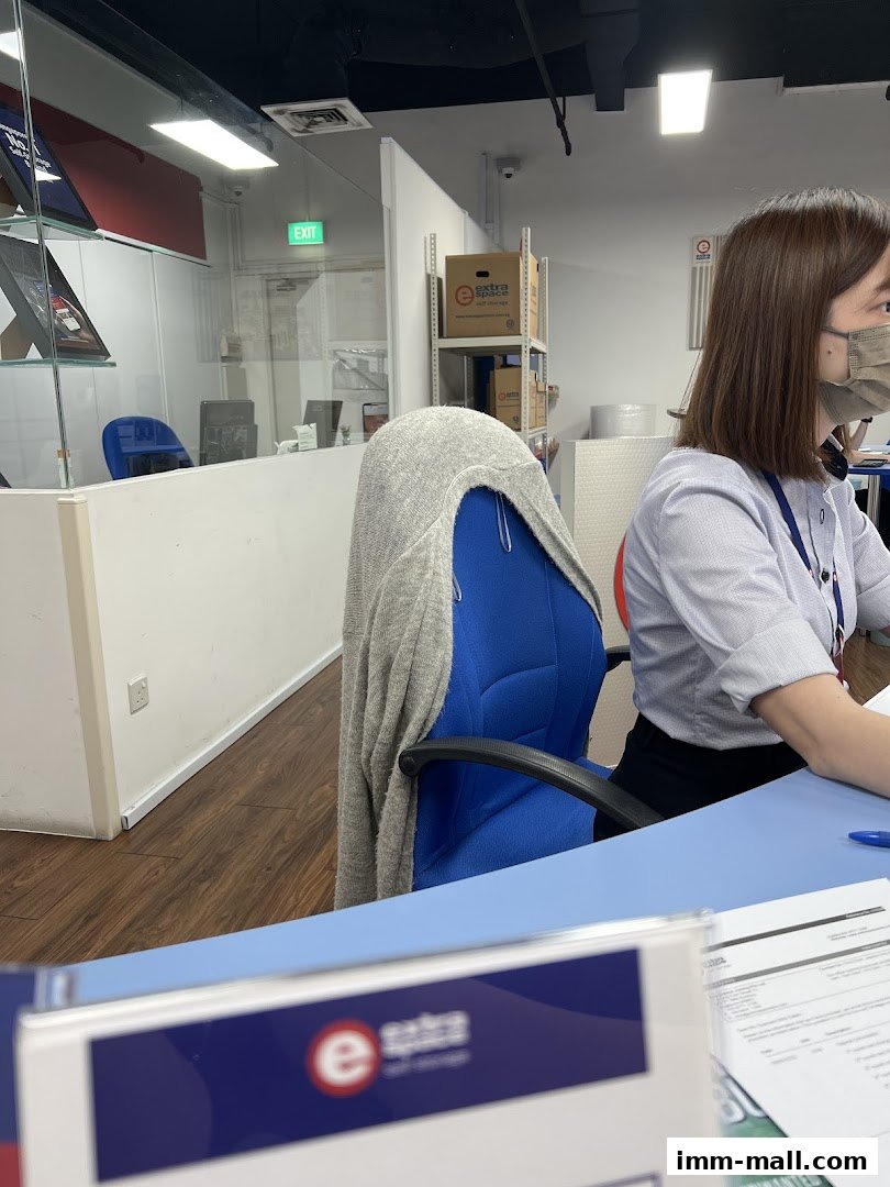 A woman wearing a mask sits at a desk in an office with storage boxes visible.