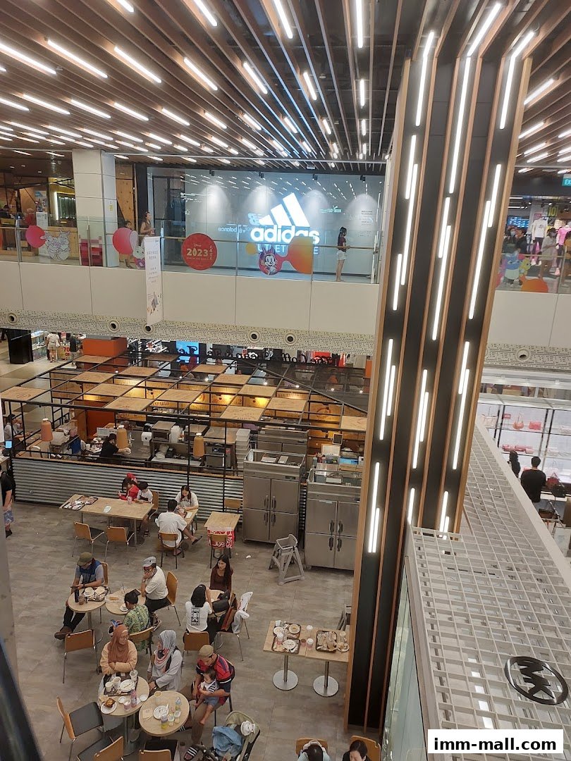A bustling indoor food court with people dining and an Adidas store visible on the upper level.