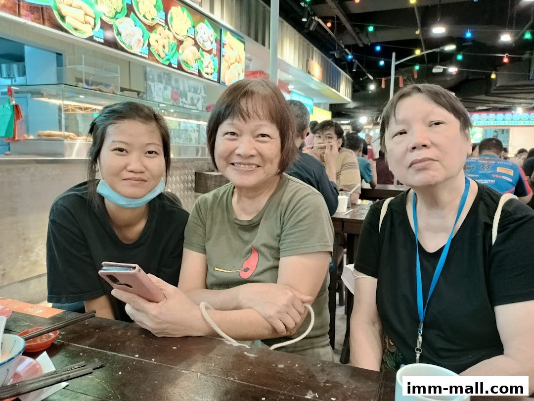 Three women are sitting at a table in a food court, with a food display in the background.
