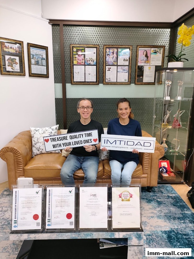 Two people sit on a couch holding signs, with framed articles and awards on the wall.