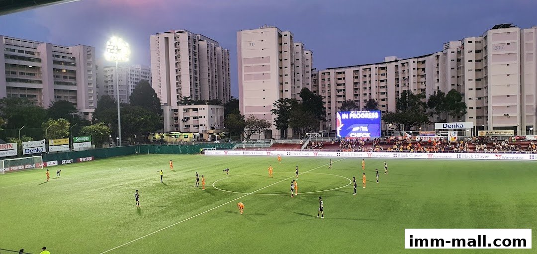 A football match is underway at Jurong Football Club stadium with players on the field and spectators in the stands.