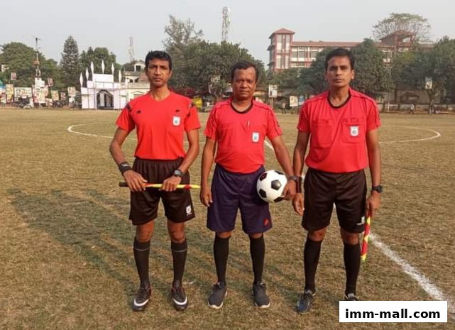 Three football referees in red shirts and black shorts stand on a grassy field with a soccer ball.
