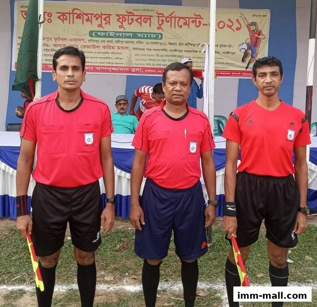 Three football referees in red shirts and shorts stand on a field with a banner behind them.