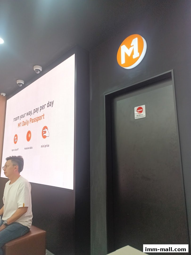 A person sits in front of a large screen displaying M1 Daily Passport promotions.