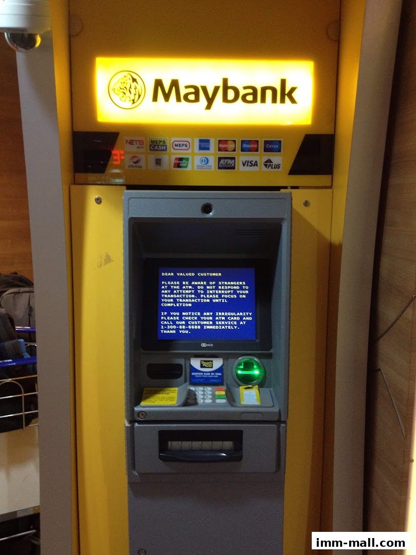 A Maybank ATM machine with various card network logos and a security message on the screen.