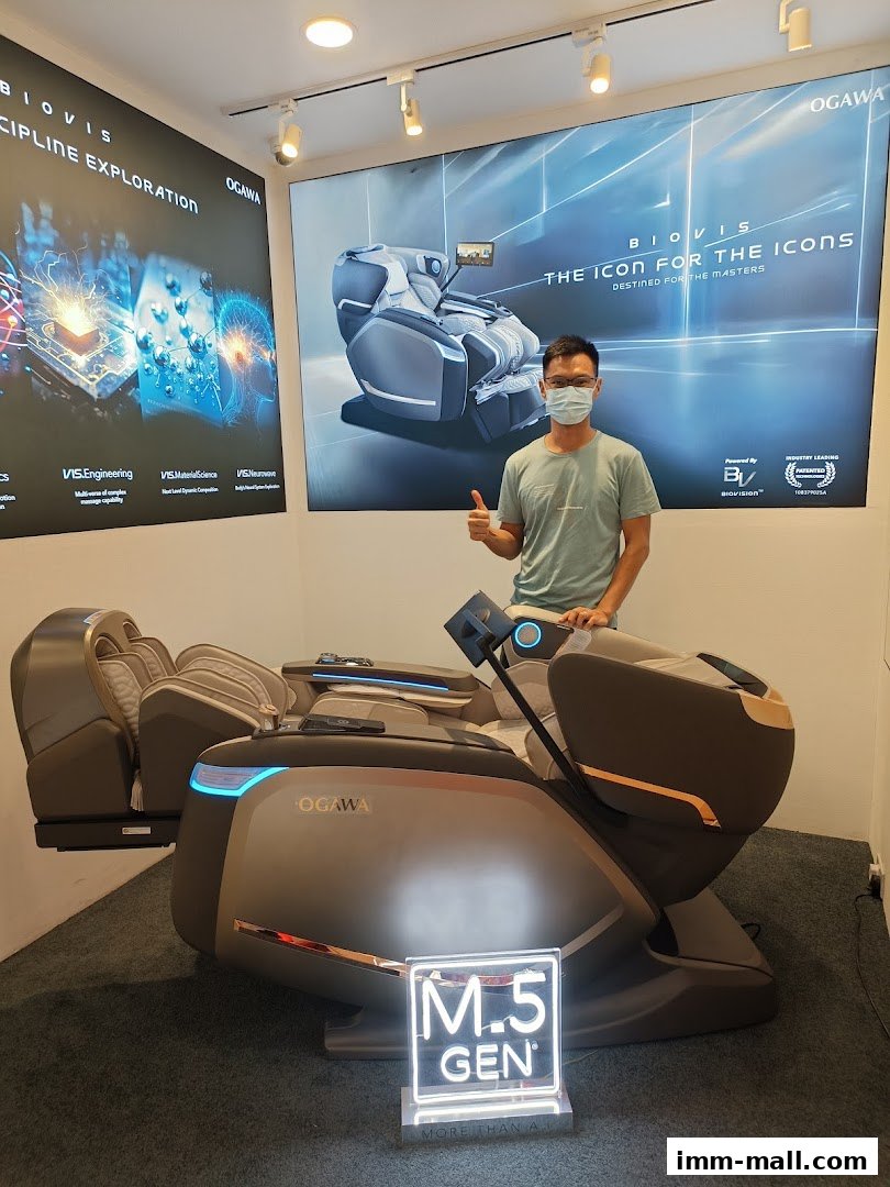 A man gives a thumbs up next to an OGAWA massage chair with promotional posters.