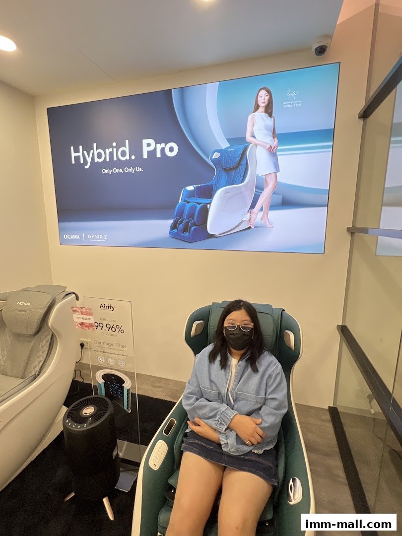 A woman sits in a massage chair in front of a large advertisement for OGAWA Hybrid. Pro.