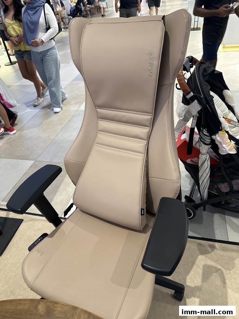A beige OSIM massage chair is displayed in a shopping mall with people walking in the background.