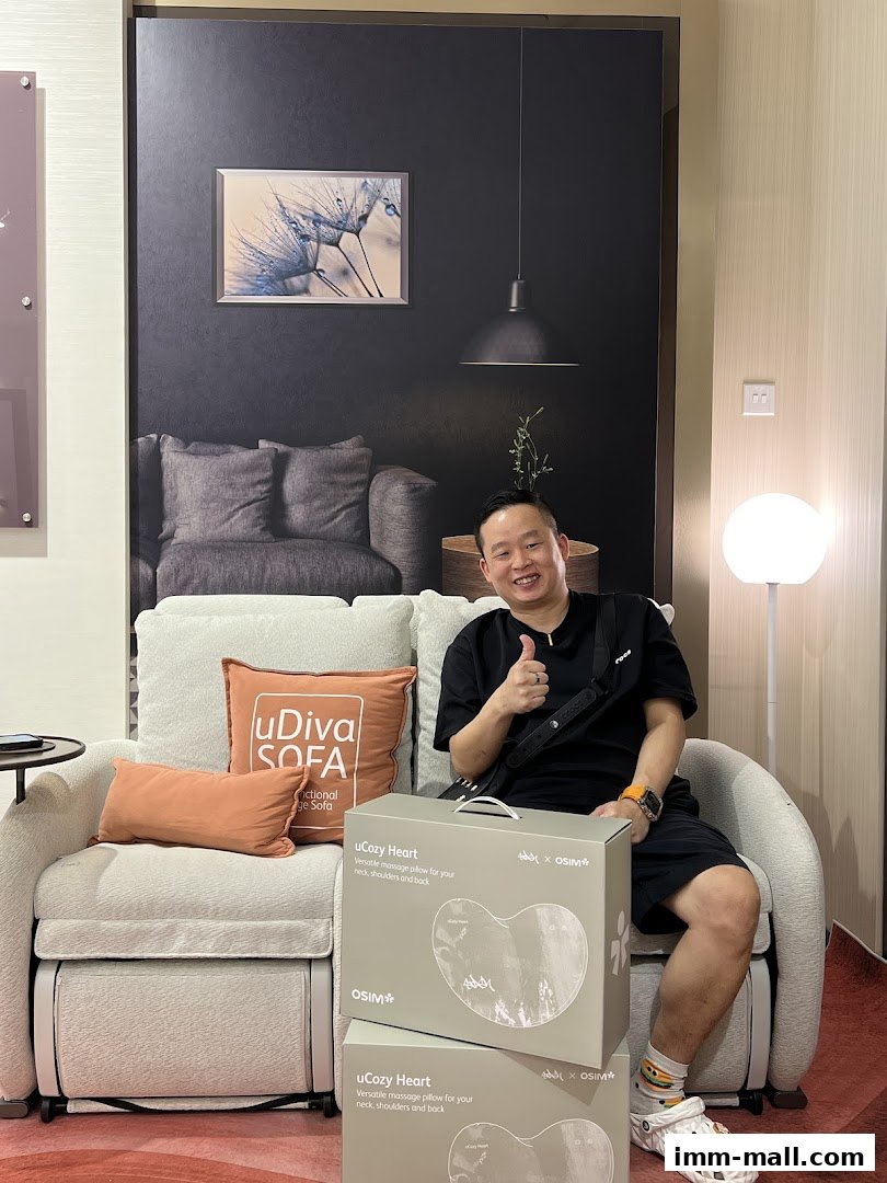 A man gives a thumbs up while sitting on a sofa with OSIM massage pillow boxes.