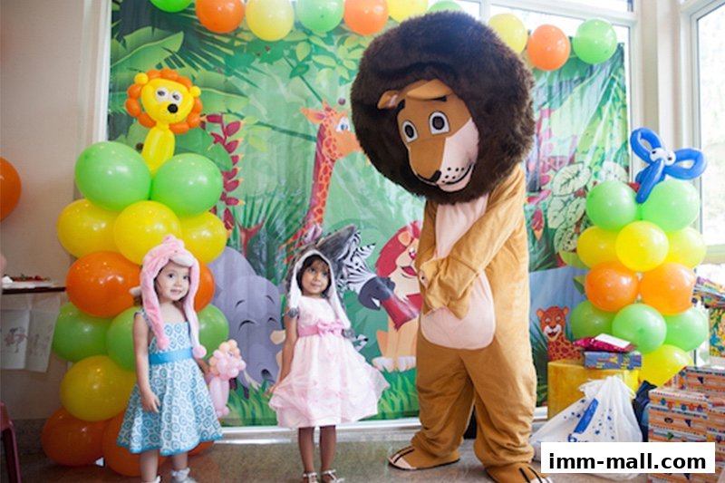 A lion mascot interacts with two young girls at a jungle-themed party with balloons.