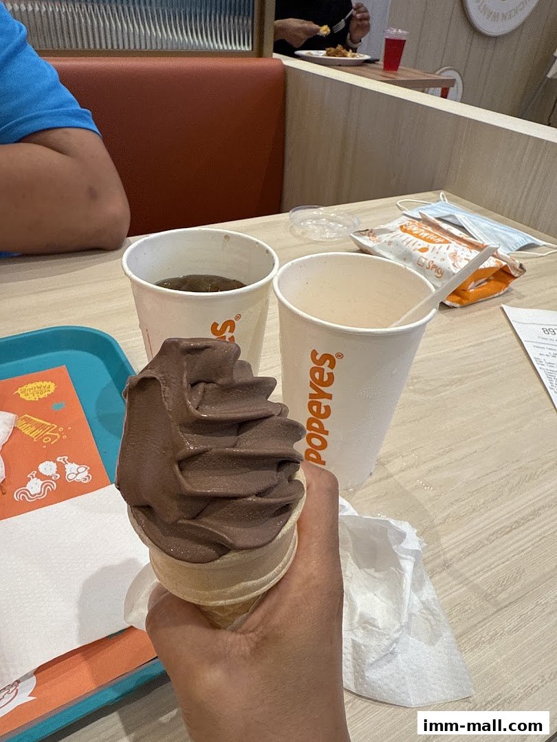 A hand holds a chocolate soft-serve ice cream cone with Popeyes cups and a bag on a table.
