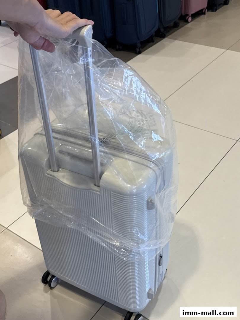 A person's hand holding the handle of a light-colored suitcase covered in plastic.