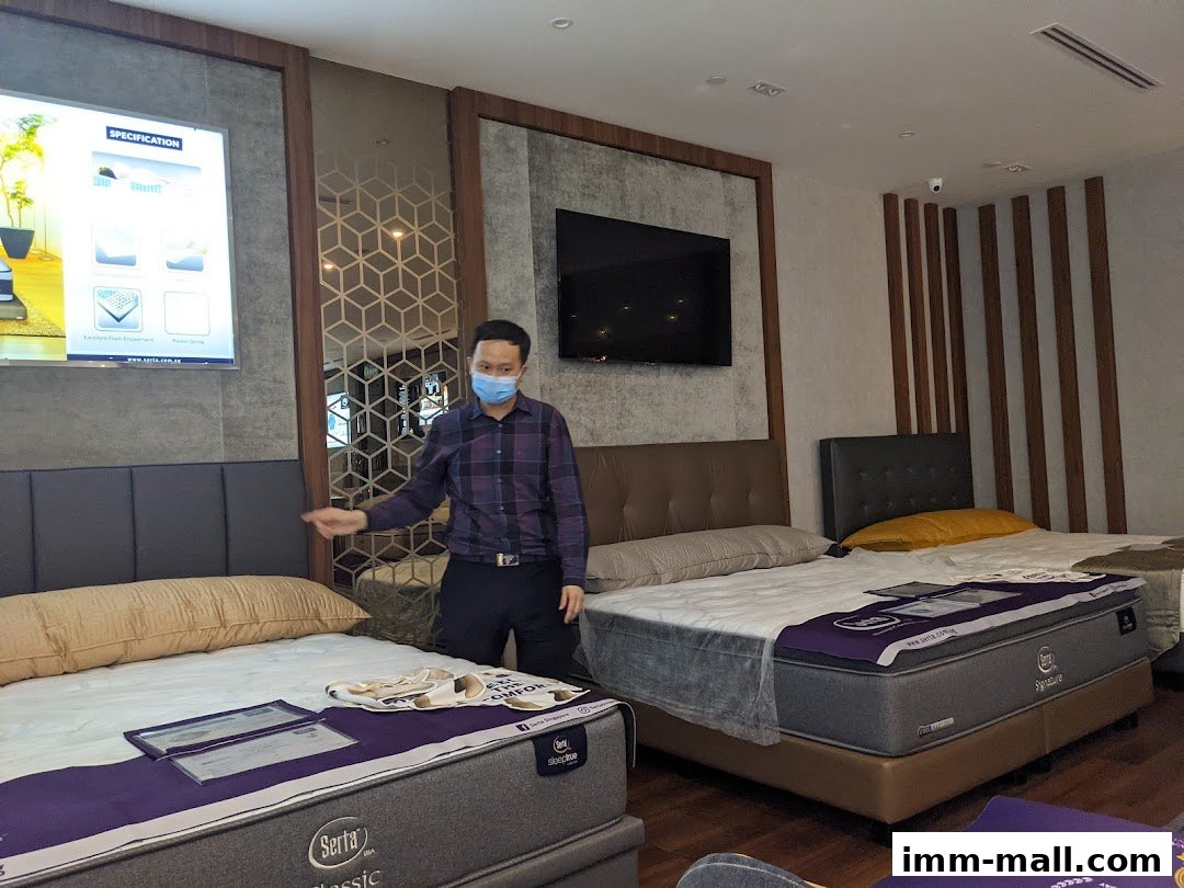 A man in a mask points to Serta mattresses displayed in a showroom.