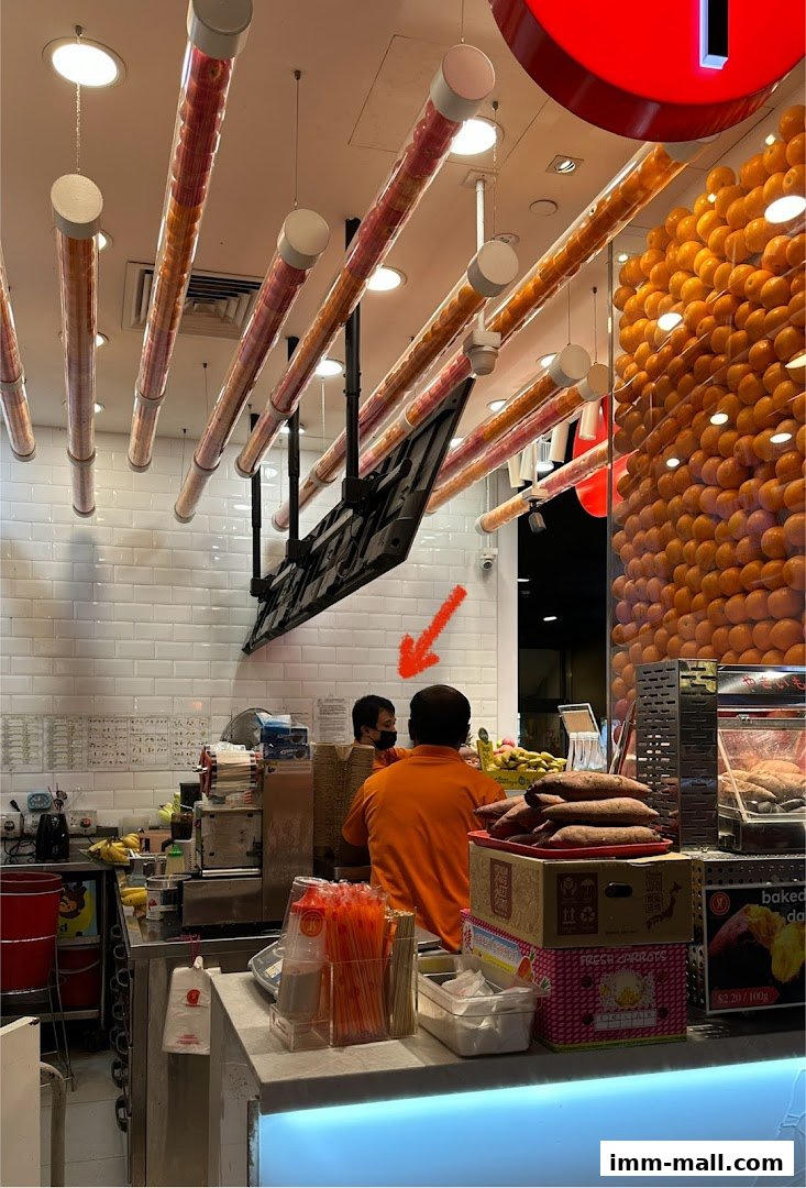 Two staff members work behind the counter at a fruit juice shop with oranges displayed.
