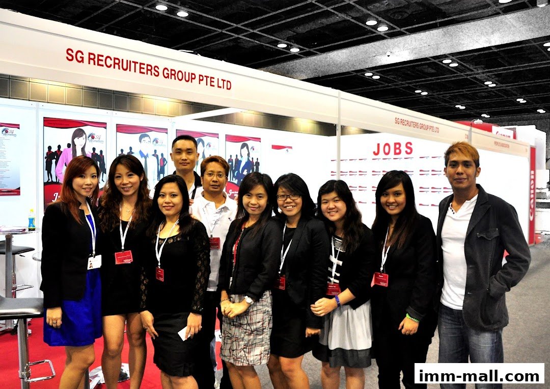 A group of smiling employees stand in front of the SG Recruiters Group Pte Ltd booth at an event.