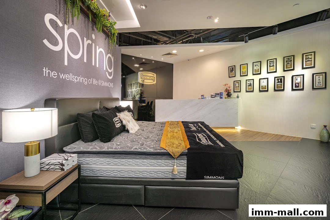 A Simmons mattress display with pillows, a lamp, and a reception desk in the background.