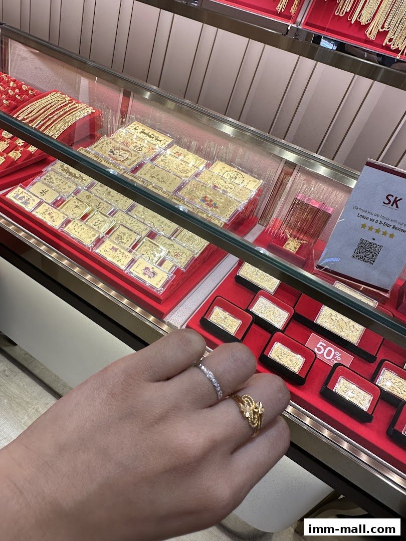 A display case filled with gold jewelry, including bars and necklaces, with a hand wearing rings in the foreground.