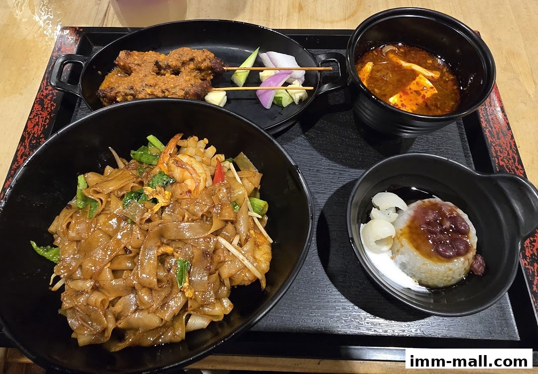 A tray of Asian food including noodles, satay, soup, and dessert.