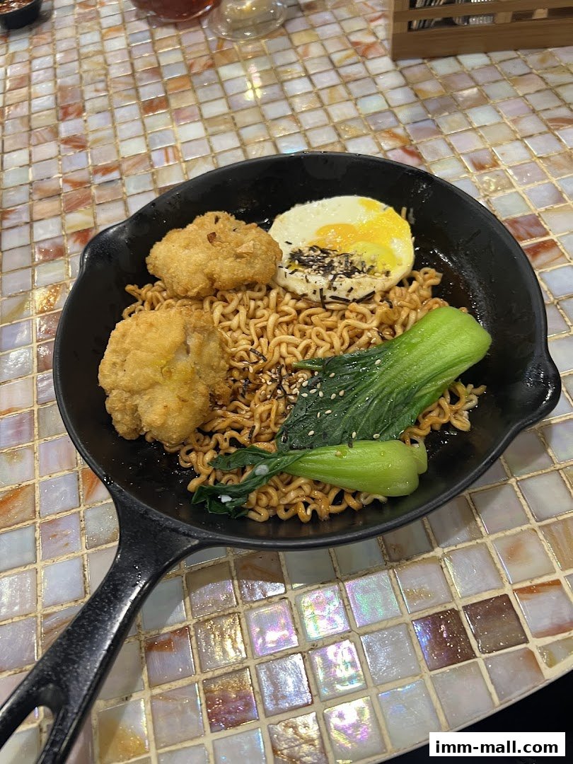 A serving of instant noodles with fried chicken, a fried egg, and bok choy in a black pan.