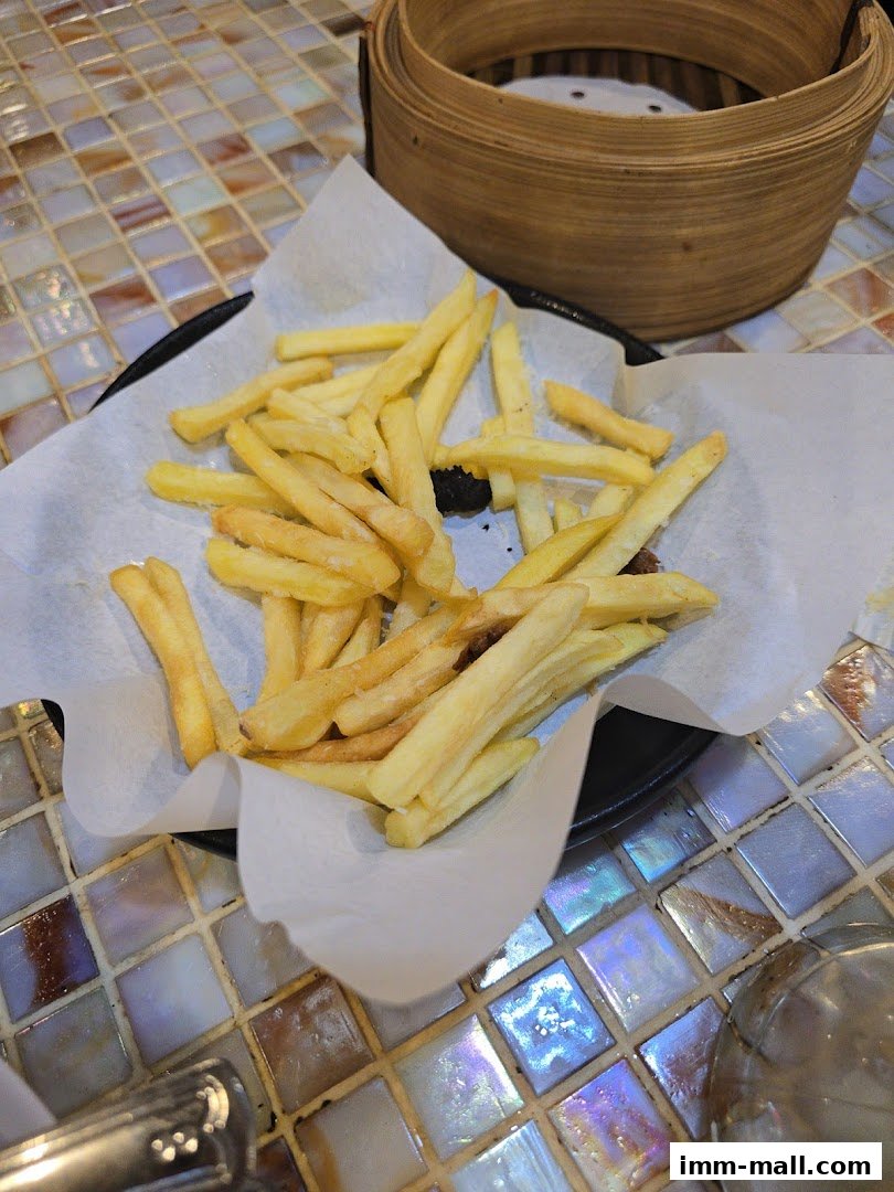 A basket of golden french fries served on white paper with a bamboo steamer in the background.