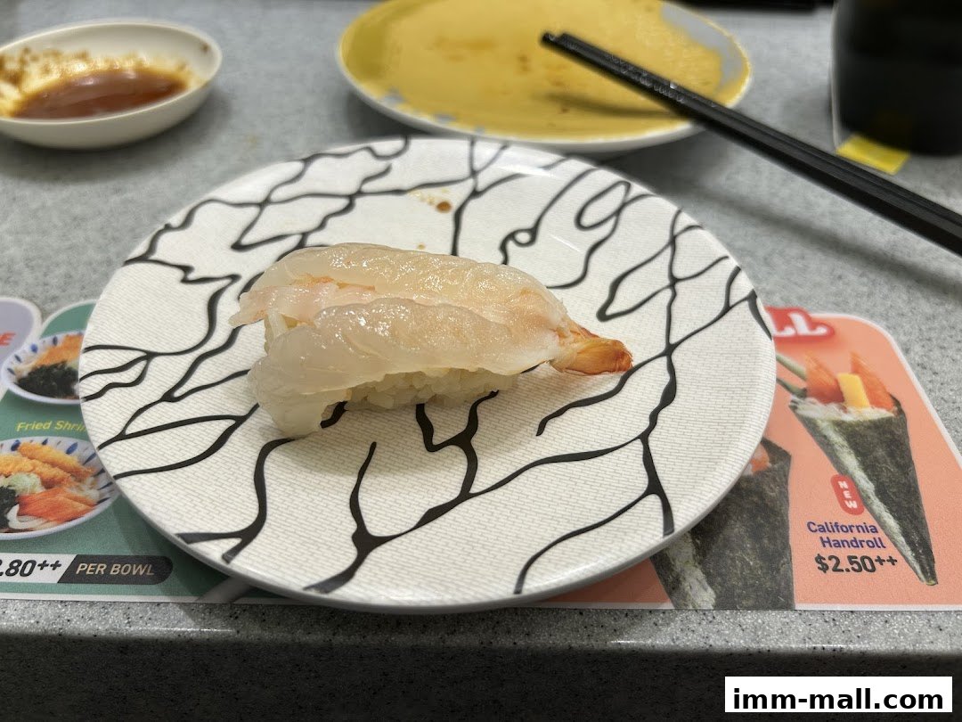 A close-up of a sushi dish with a shrimp topping on a patterned plate, with other food items in the background.