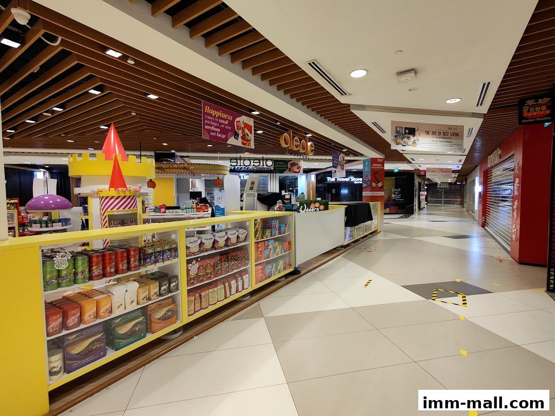 A brightly lit shopping mall corridor with various shops and displays, including a yellow stall with packaged goods.