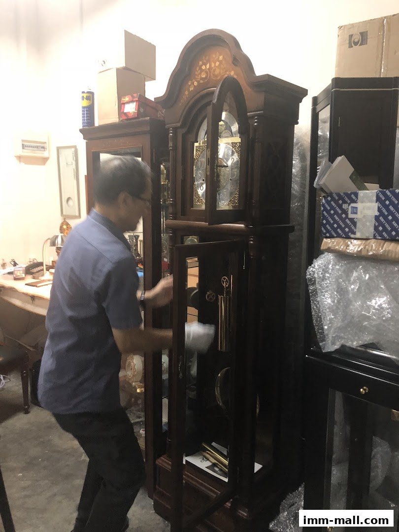 A man is cleaning a tall grandfather clock in a shop with other items in the background.