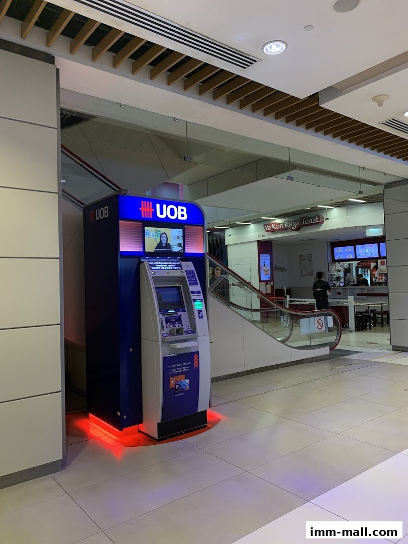 A UOB ATM machine is located in a shopping mall, with an escalator and food stalls in the background.