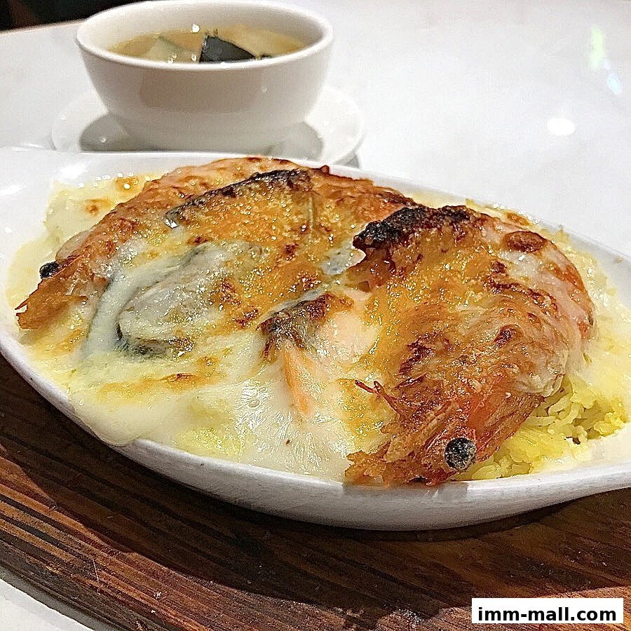 A close-up of a baked dish with cheese, shrimp, and rice, served with a side of soup.
