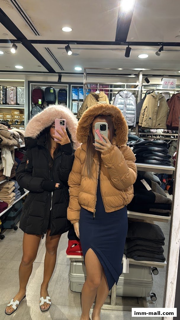 Two women in puffer jackets with fur hoods pose for a mirror selfie in a clothing store.